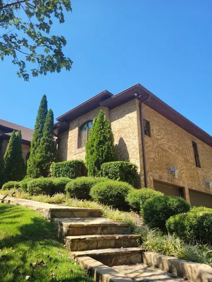 A large brick house with a brown roof, surrounded by green bushes and trees.